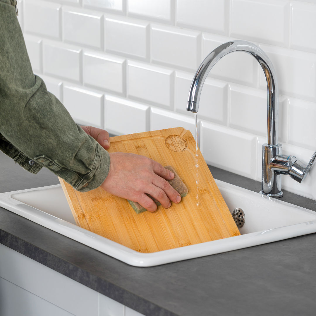 Person cleaning a wooden cutting board in a kitchen sink with a sponge.